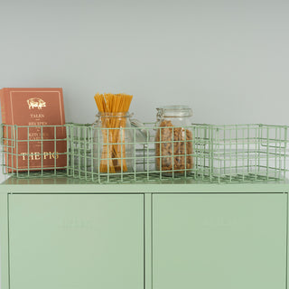 A set of three sage green metal baskets containing a book and two jars, on top of a sage green storage locker.